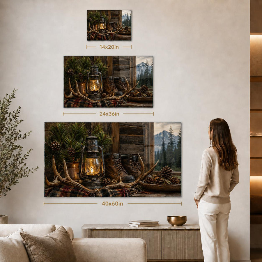 A woman stands in a living room beside a wall displaying three rustic wall art prints with overlaid size labels: 14x20in, 24x36in, and 40x60in. The prints show a forest cabin scene with antlers, lanterns, pinecones, and mountains.